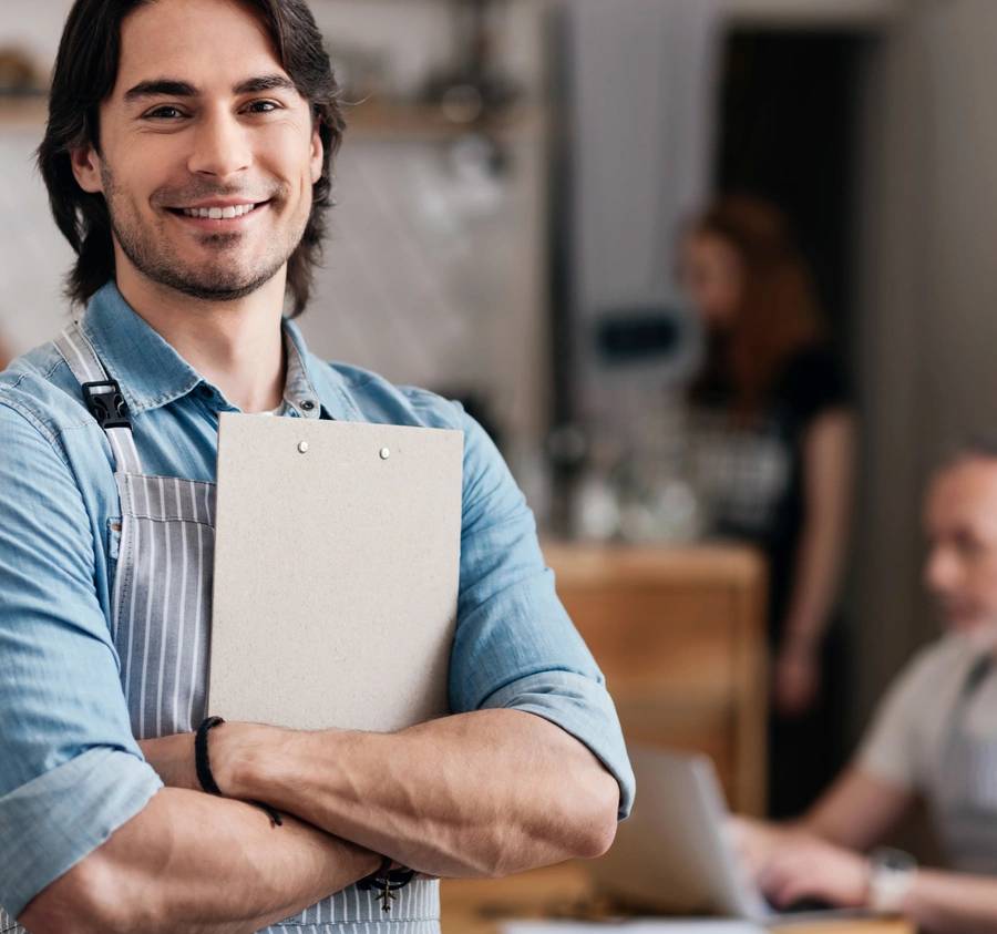 Handsome diner. Cheerful and positive male cafe worker holding a menu while standing and happily smiling at a camera with some people in a background (Handsome diner. Cheerful and positive male cafe worker holding a menu while standing and happily smi